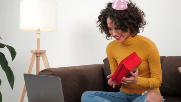 Happy woman in party hat opens gift box and surprised, sitting in couch at home — ストック動画