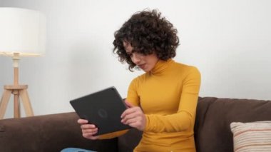 Curly woman emotionally playing video game on tablet, sitting on couch at home