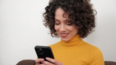 Gülümseyen kıvırcık kadın akıllı telefon dinleme ekranı kullanıyor, evdeki kanepede oturuyor.