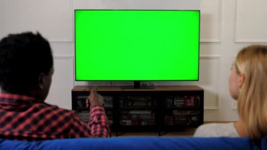 Yeşil ekran TV 'ye bakan çok ırklı çift uzaktan kumandada kanal değiştiriyor