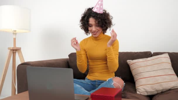 Happy woman in party hat opens gift box and surprised, sitting in couch at home — ストック動画