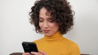 Smiling curly woman uses smartphone tapping screen display, sitting on couch at home. Positive businesswoman works for mobile phone online chatting with company employees, distance business meeting