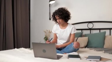 Kıvırcık kadın öğrencilerin online video çalışması webcam laptop 'u arıyor not defterine bilgi yazıyor, uzaktan eğitim. İspanyol kadın bilgisayardaki yüksek lisans dersini seyrediyor evde yatakta oturuyor.