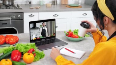 Bilgisayar ekranındaki erkek yemek blogcusu ev hanımlarına online video kameralı laptop öğretiyor, domates dilimliyor. Mutfaktaki kadın uzaktan aşçılık dersi alıyor. Şef öğretmenin kahve içmesini dinle.