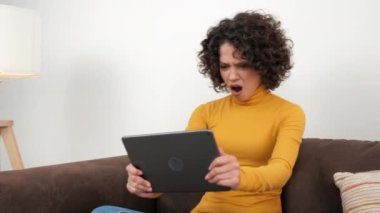 Hispanic curly woman gamer emotionally playing video game on tablet, sitting on couch at home. Positive Latin American female intensively plays racing games on the tablet, wearing yellow turtleneck