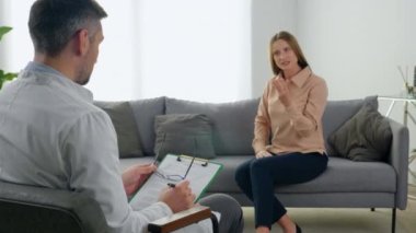 Psikoterapist adam hastayı dinler ve psikoterapi seansında not alır. Zihinsel sorunları olan sinirli ve odaklanmış bir kadın koltukta oturmuş terapistle konuşuyor.