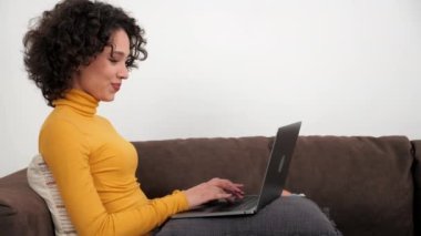 Gülümseyen iş kadını evdeki şirket çalışanlarıyla çevrimiçi olarak sohbet ediyor. Pozitif İspanyol kıvırcık kadın öğrenci çalışması klavyede dizüstü bilgisayar kullanıyor. Koltukta oturuyor, uzaktan eğitim alıyor.