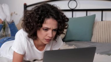 Odaklanmış İspanyol kıvırcık kadın öğrenci çalışması dizüstü bilgisayarını kullanarak yatağa uzanır, uzaktan eğitim alır. Konsantre iş kadını evde çalışan şirket çalışanlarıyla çevrimiçi olarak sohbet ediyor.