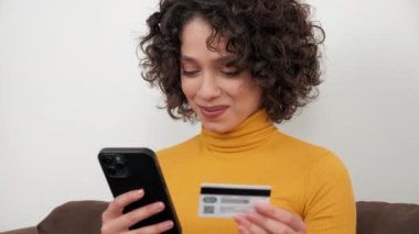 Gülümseyen kıvırcık kadın müşteri hediye ödemek için kredi kartı koduna giriyor, akıllı telefondan online alışveriş yapıyor. Mutlu kadın alışverişçiler internet alışverişleri ya da cep telefonuyla yapılan ödemeler. Çevrimiçi bankacılık