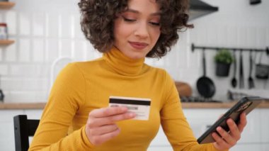 Close up curly woman customer enters credit card code to pay gifts, online shopping via smartphone. Female shopper internet purchases or payments via mobile phone at home kitchen. Online banking