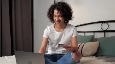 Gülümseyen kıvırcık kadın müşteri hediye ödemek için kredi kartı koduna giriyor, bilgisayarla online alışveriş yapıyor. Mutlu kadın alışverişçiler internet alışverişleri ya da evdeki bilgisayarla ödemeler, yatakta oturmalar. Çevrimiçi bankacılık