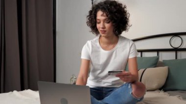Gülümseyen kıvırcık kadın müşteri hediye ödemek için kredi kartı koduna giriyor, bilgisayarla online alışveriş yapıyor. Mutlu kadın alışverişçiler internet alışverişleri ya da evdeki bilgisayarla ödemeler, yatakta oturmalar. Çevrimiçi bankacılık