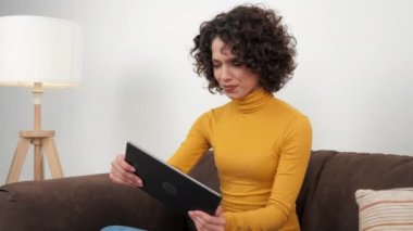 Hispanic curly woman gamer emotionally playing video game on tablet, sitting on couch at home. Positive Latin American female intensively plays racing games on the tablet, wearing yellow turtleneck