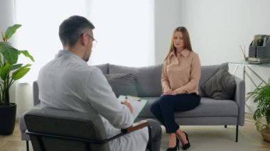 Sad adult woman with mental health problems after covid-19 sitting on couch talking to therapist. Man doctor psychotherapist listen patient at psychotherapy session and taking notes on clipboard