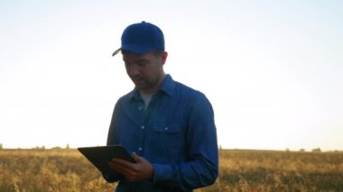 Smiling farmer businessman is working on tablet computer standing in wheat field at sunrise. Positive man uses tablet tapping screen, plans to harvest. Concept technology in agriculture, farming