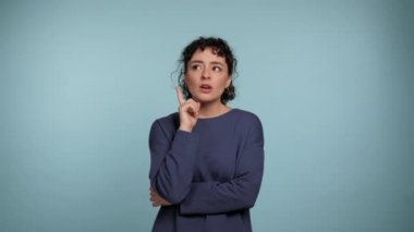 Pensive young curly woman in blue sweater posing looking around, thinking raising finger with idea. Thoughtful beautiful female thinks how to solve the problem on isolated light blue background