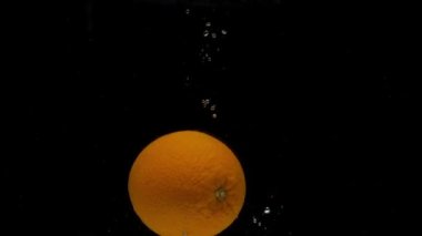 Ağır çekimde bir portakal siyah arkaplanda şeffaf suya düşüyor. Akvaryuma taze narenciye sıçrıyor. Meyve, yemek.