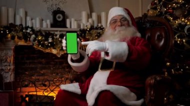 Modern gülümseyen Noel Baba akıllı telefon yeşil ekranını işaret ediyor. Pozitif Noel Baba elini cep telefonuyla gösteriyor. Renkli anahtar görüntüsü, Noel alışverişi, yeni yıl hediyeleri, tatiller.