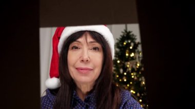 View from inside carton box aged woman in Santa Claus hat opens Christmas gift box and surprised. Smiling female opens New Year gift parcel on background Christmas tree, celebration Christmas holidays