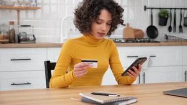 Smiling curly woman customer enters credit card code to pay gifts, online shopping via smartphone. Female shopper internet purchases or payments via mobile phone at home kitchen. Online banking