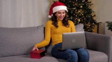 Noel Baba şapkalı kadın kırmızı hediye kutusunu açıyor ve şaşırıyor. Şok olmuş bayanların online kutlama videosu arka plandaki koltukta oturuyor Noel ağacı, Noel tatili, mutlu yıllar