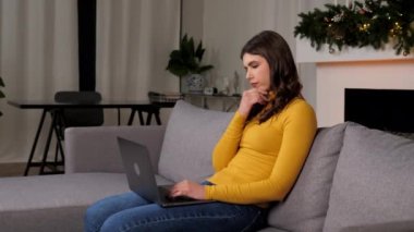 Thoughtful businesswoman uses laptop distance works for computer typing on keyboard sitting on couch at cozy home. Thinking woman online chatting with best friends. Christmas, New Year winter holidays