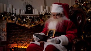 Modern gülümseyen Noel Baba oyuncusu arka plandaki şöminede, Noel ağacında mobil oyun oynuyor. Yaşlı büyükbaba Noel Baba kostümü giymiş. Tablet kullanıyor. Sandalyeye oturuyor. Yeni yıl tatili