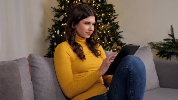 笑顔の女性は バックグラウンドクリスマスツリー上のタブレットを使用してインターネット上で新年の贈り物を購入します ポジティブ女性は居心地の良い家でソファに座ってオンラインチャットポータブルデバイスを使用します 冬休み — ストック動画