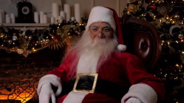 Gülümseyen Noel Baba arka planda yanan şöminenin üzerinde oturan kameraya bakar, Noel ağacı. Noel Baba kostümü giymiş yaşlı bir adam. Yeni yılın kutlu olsun. 