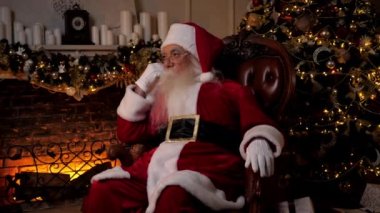Düşünceli Noel Baba, arka planda yanan şöminenin üzerinde oturan sandalyeye bakıyor, Noel ağacı. Noel Baba kostümü giymiş dalgın yaşlı bir dede. Yeni yılın kutlu olsun.