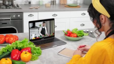 Mutfaktaki kadın uzaktan aşçılık dersi alıyor şef öğretmeni dinliyor, defterine yazıyor. Bilgisayar ekranındaki erkek yemek blogcusu ev hanımına online video kameralı laptop dersi veriyor, salatalığı dilimliyor