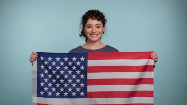 アメリカ国旗を掲げた青いセーターを着た若い巻き毛の女性が笑顔でカメラのように見える 孤立したライトブルーの背景にアメリカのフラグを持つ肖像画の美しい女性立っている 7月4日アメリカ独立記念日 — ストック動画
