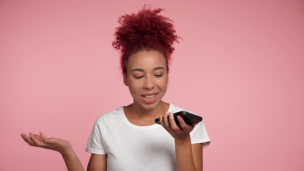 笑顔を閉じるアフリカ系アメリカ人の赤毛の巻き女性はスマートフォンを使用して友人にオーディオメッセージを指示します 肖像画正幸せな女性の話上の携帯電話上の隔離されたピンクの背景 — ストック動画
