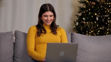 Gülümseyen genç bir kadın arka plandaki Noel ağacında bilgisayarla en iyi arkadaşlarıyla sohbet ediyor. Pozitif güzel kadın mesafesi evdeki kanepede oturan klavyedeki bilgisayar daktilo metninde işe yarar.