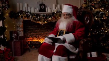 Modern Noel Baba tablet üzerinde çalışır. İnternetten çocuklara Noel hediyesi alır. Arka plandaki şöminede, Noel ağacında, mutlu yıllar. Adam tablet kullanıyor. İnternetten alışveriş yapıyor. Sandalyede oturuyor.