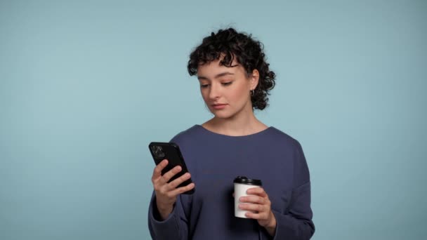 严重的卷曲女商人使用智能手机打字浏览孤立的浅蓝色背景 漂亮的女人拿着手机上网远程工作 手里拿着咖啡或茶 — 图库视频影像