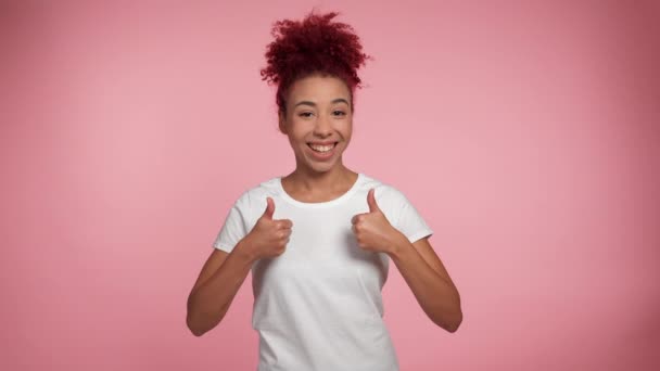 アフリカ系アメリカ人の赤毛の巻き毛の女性が親指を上げるような手のショーを笑顔肖像画はカメラに見えます コピースペース付きの隔離されたピンクの背景の白いTシャツの喜びの女性 人々の感情 — ストック動画