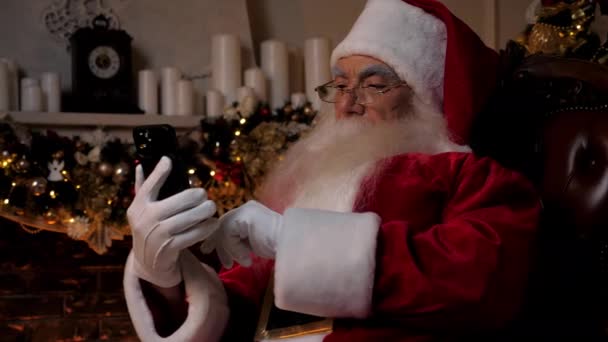 在智能手机上工作的现代圣诞老人在网上为孩子们买圣诞礼物 新年快乐 身穿圣诞老人装的爷爷坐在椅子上 用智能手机在网上购物 — 图库视频影像