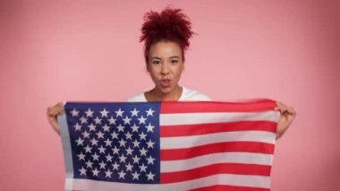 Gülümseyen Afro-Amerikan vatansever kızıl saçlı, Amerikan bayrağı tutan kadın, ABD 'nin kameraya benzediğini söylüyor. Amerikan bayrağıyla izole edilmiş pembe arka planda duran portre kadın. 4 Temmuz Amerika Bağımsızlık Günü