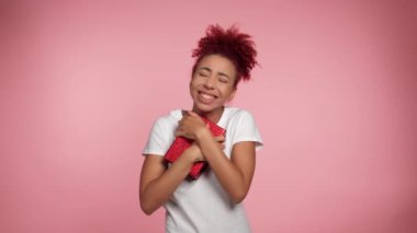 Mutlu Afro-Amerikan kıvırcık saçlı kadın doğum günü hediyesini kucaklıyor. Kırmızı kutu içinde ne olduğunu merak ediyor. İzole edilmiş pembe arka planda elinde hediyeyle beyaz tişörtlü, gülümseyen güzel bir kadın.