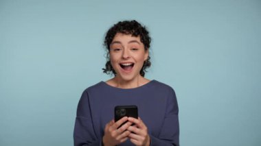 Akıllı telefon kullanarak gülümseyen kıvırcık kadın izole açık mavi arka planda vay canına diyor. Mutlu pozitif kadın cep telefonu satışı büyük bir şok yarattı. İnsanların duyguları yaşam tarzı