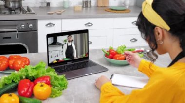 Mutfaktaki kadın video çalışıyor webcam laptop arıyor şef öğretmeni dinliyor, deftere yazıyor. Bilgisayar ekranındaki adam yemek blogcusu ev hanımına uzaktan aşçılık dersi veriyor, domates dilimliyor