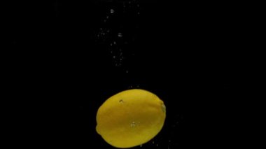 Yavaş çekimde bir sarı limon siyah arkaplanda şeffaf suya düşüyor. Akvaryuma taze meyve sıçraması. Citrus, yemek, hava kabarcıkları