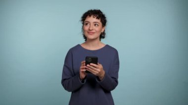 Gülümseyen kıvırcık kadın akıllı telefon kullanıyor. İnternetten alışveriş yapmayı düşünüyor. Cep telefonlu kadın izole edilmiş açık mavi arka planda online satın alma kararı aldı