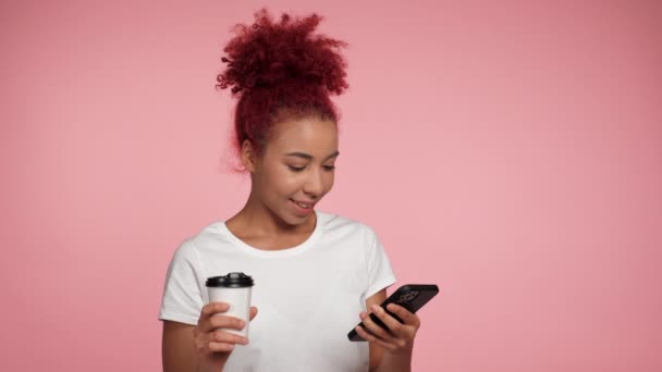 近距离观察微笑的非洲裔美国女商人 用打字浏览智能手机 看着相机 形象红头发卷曲的女人在孤立的粉色背景下的在线遥控器上工作 — 图库视频影像
