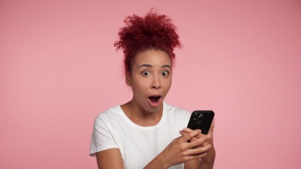 Schockierte Afroamerikanische Rothaarige Lockenkopf Frau Mit Smartphone Auf Isoliertem Rosa — Stockvideo