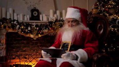Modern gülümseyen Noel Baba arka plandaki şöminede, Noel ağacında mobil oyun oynar. Yaşlı büyükbaba Noel Baba kostümü giymiş. Tablet kullanıyor. Sandalyeye oturuyor. Yeni yıl kış tatili
