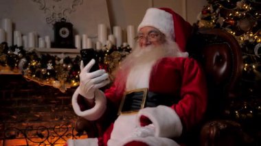 Modern gülen Noel Baba akıllı telefondan komik şovlar izliyor. Dedem Noel Baba kostümü giymiş. Akıllı telefon kullanıyor. İnternette komik videolar izliyor. Mutlu yıllar, Noel