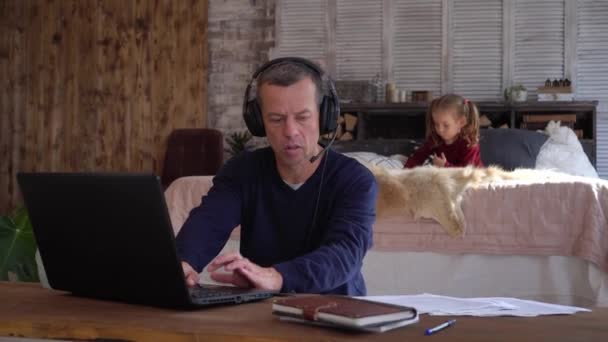 En man fjärrkontroll arbetar med ett litet barn i hemmakontoret under Covid-19 nedstängning. Företagsvideokonferens. Pappa jobbar i vardagsrummet med hörlurar och mikrofon. Frilansjobb online — Stockvideo