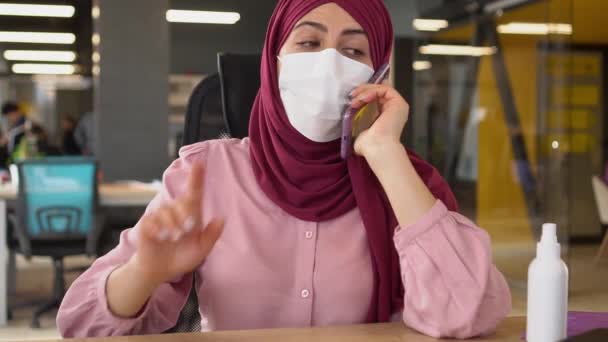 穿着头巾的现代穆斯林女商人在办公室里工作，在电话里聊天。COVID-19大流行病。工人和客户的健康工作场所。医疗面罩、手部清洁剂和社交距离 — 图库视频影像